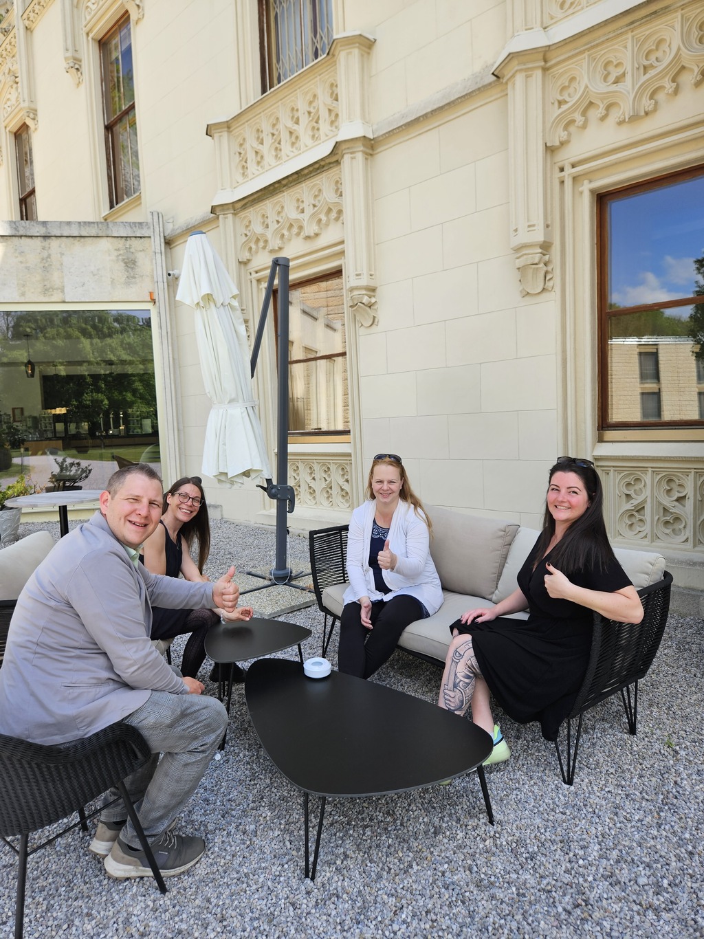 Vier Teammitglieder der Seewald Services GmbH sitzen entspannt im Freien vor einem historischen Gebäude, lächeln in die Kamera und zeigen Daumen hoch.