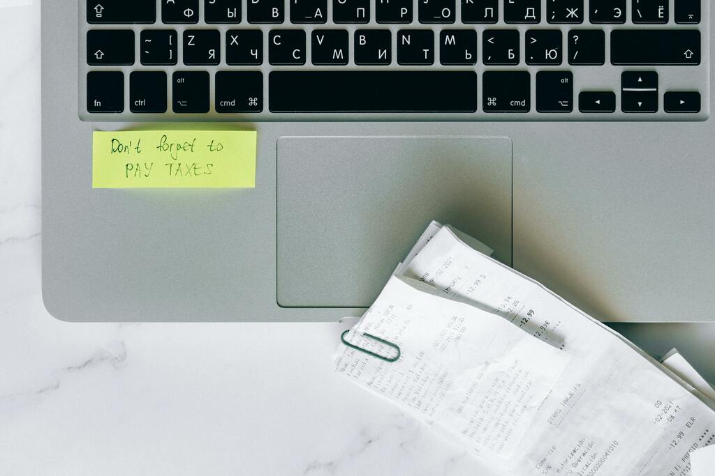 Laptop-Tastatur mit gelbem Post-it und gestapelten Dokumenten auf Schreibtisch, Symbolbild für Büroservice.