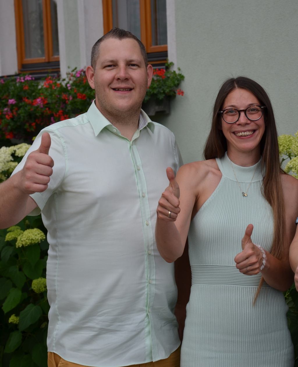 Dominik Seewald und Petra Seewald, MSc, vor einem Gebäude mit Blumen im Hintergrund, zeigen Daumen hoch und freuen sich auf deine Bewerbung.